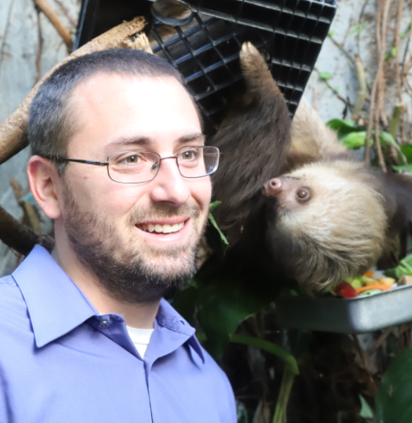 smiling male with glasses with standing next to brown sloth